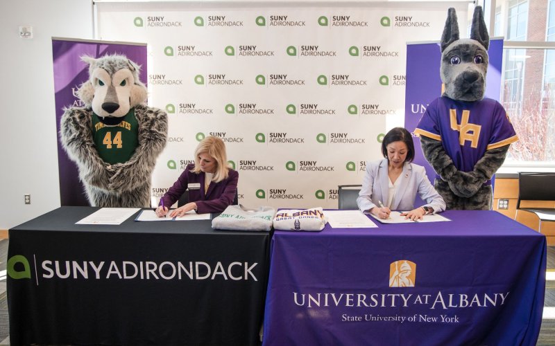 Two women sign documents at tables with tablecloths saying SUNY Adirondack and Univeristy at Albany, as mascots of a timberwolf and a great dane stand on either side