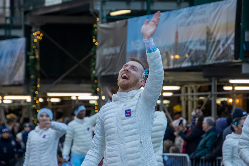 A man in a white coat smiles and waves in a crowd in New York City.