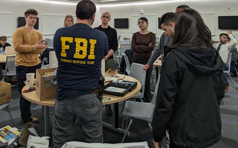 The back of a man is seen wearing a T-shirt that reads "FBI Evidence Response Team" standing in front of a round table surrounded by students. 