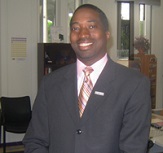 Ekow King in a gray suit with a gold tie smiling and standing in front of campus center windows.