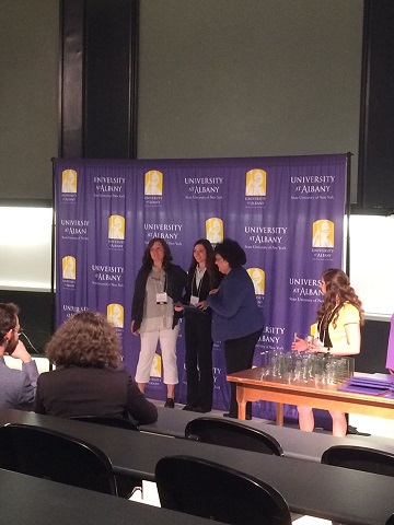 Three women stand at the front of a conference room in front of a UAlbany backdrop. The person in the middle holds a plaque.