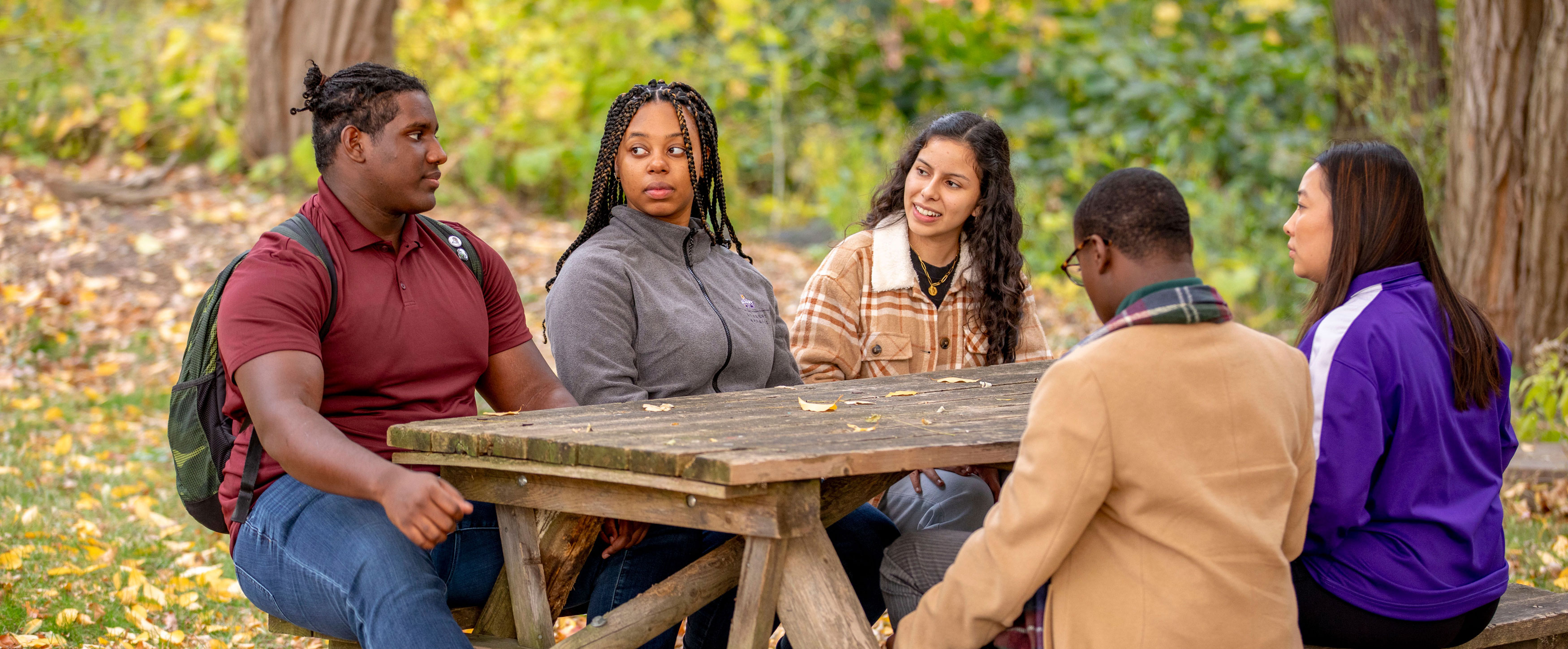 Five students sit and talk at a picnic table outside under trees with fall foliage.