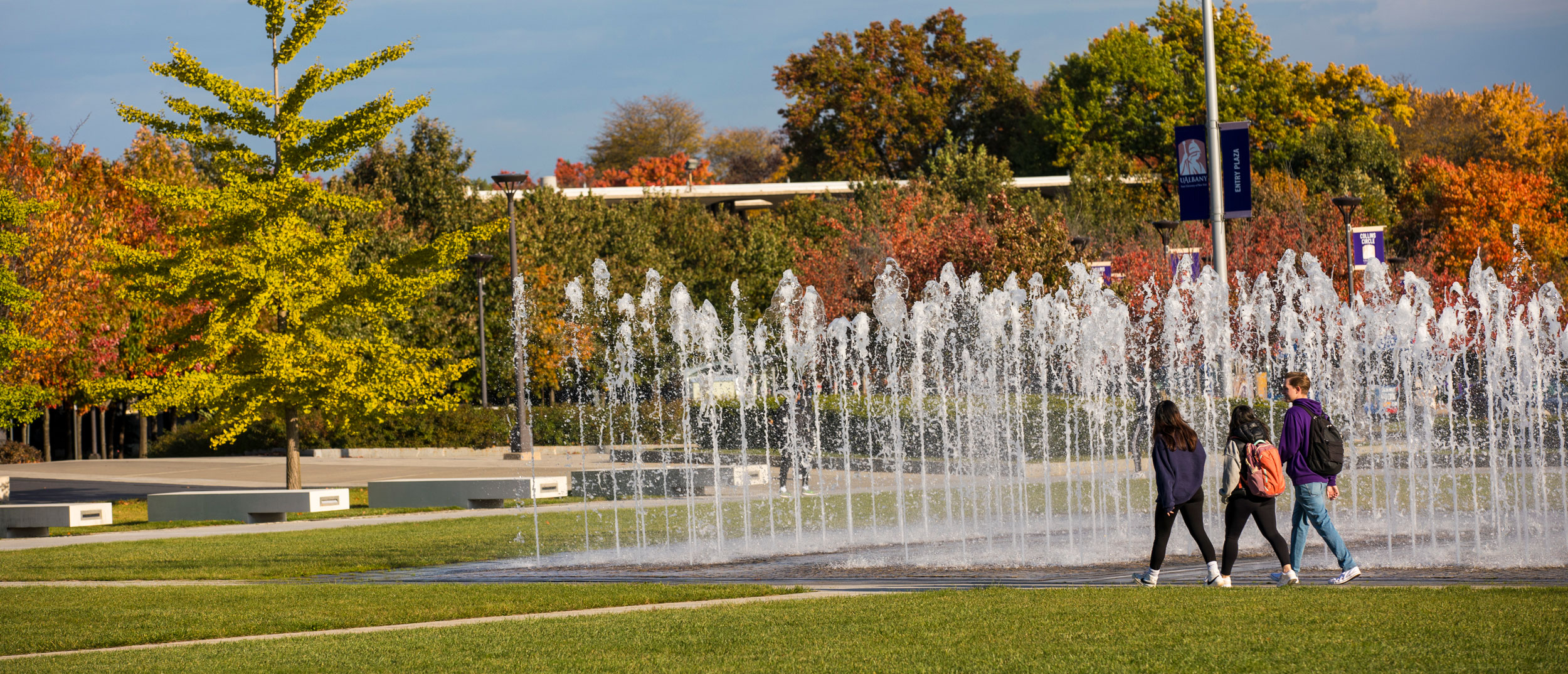 Three students with backpacks walk along a campus pathway next to a fountain.