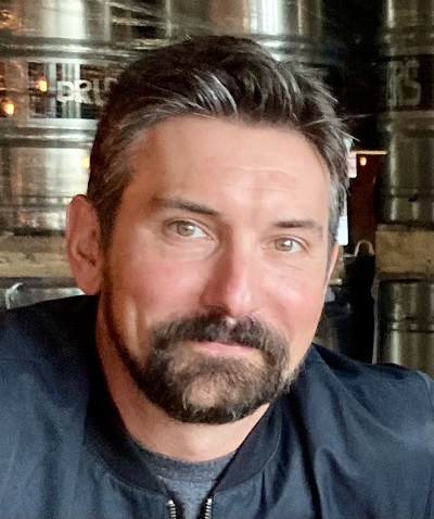 A man with short brown and gray hair and a brown beard smiles for a photo.