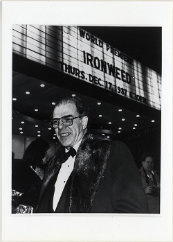 A man in glasses and a tuxedo and overcoat stands smiling in front of an illuminated theater marquee.