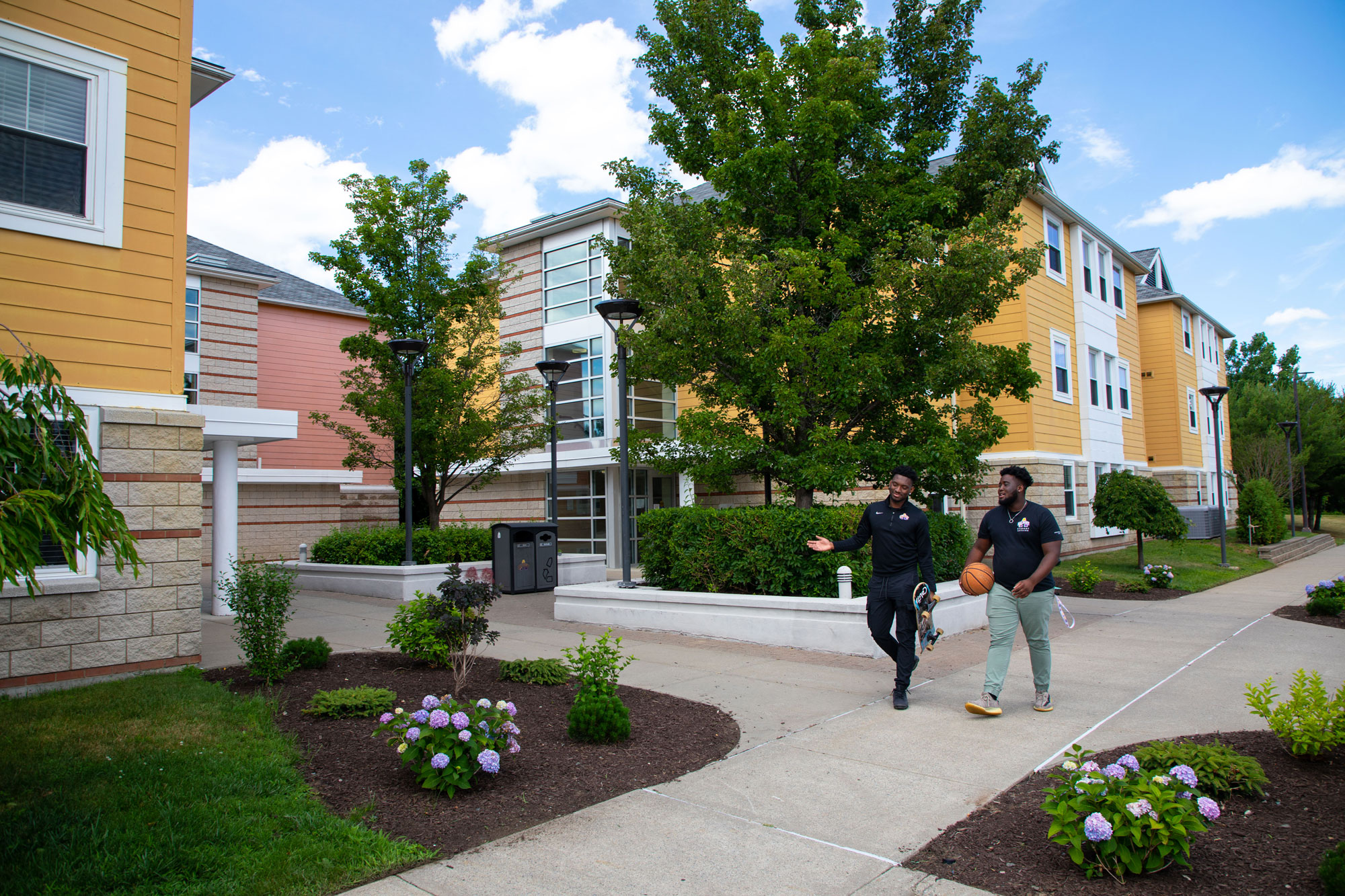 Two students with a basketball walk through Empire Commons, an apartment complex. The buildings are pink and yellow, with green trees and grass.