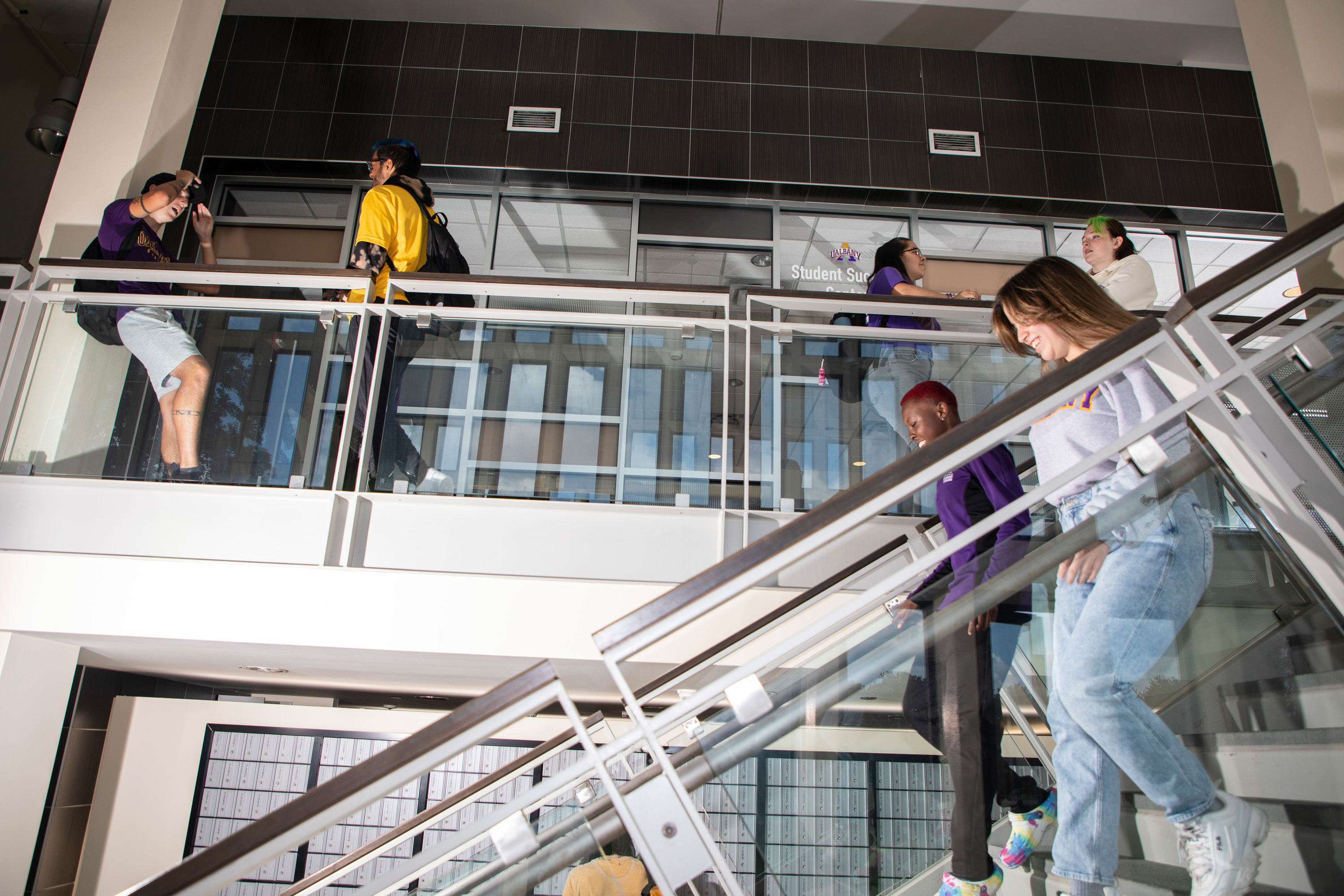 Four students stand and talk on an upper level inside State Quad, while two other students walk down a staircase outside the Student Success Center. The atrium is built with gray stone and glass.