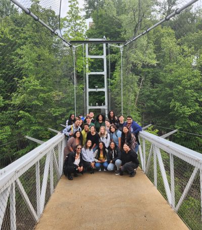 A group of students gather on a narrow suspension bridge with leafy trees seen behind them