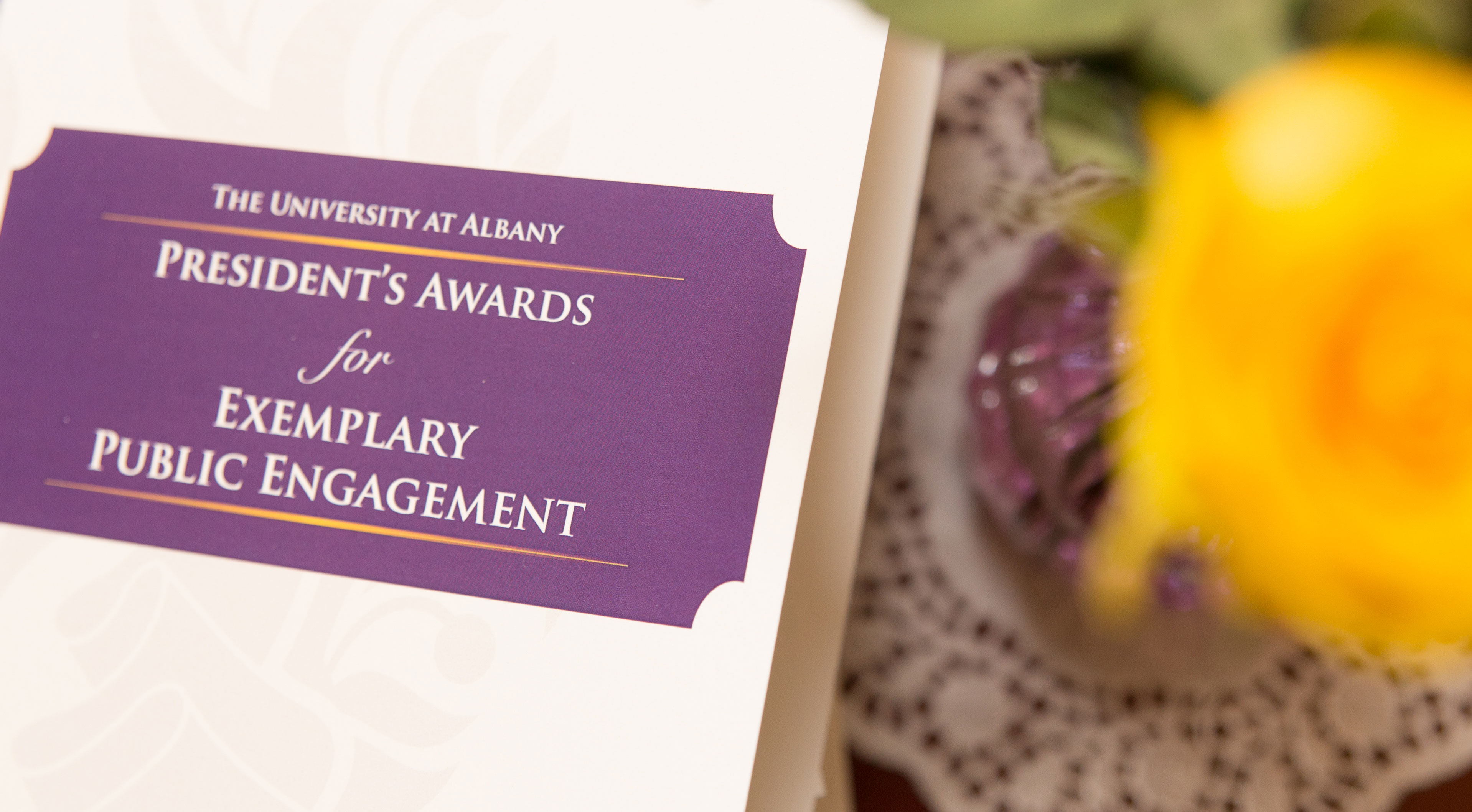 A white and purple program with the words "The University at Albany President's Awards for Exemplary Public Engagement" sits beside a yellow rose in a vase