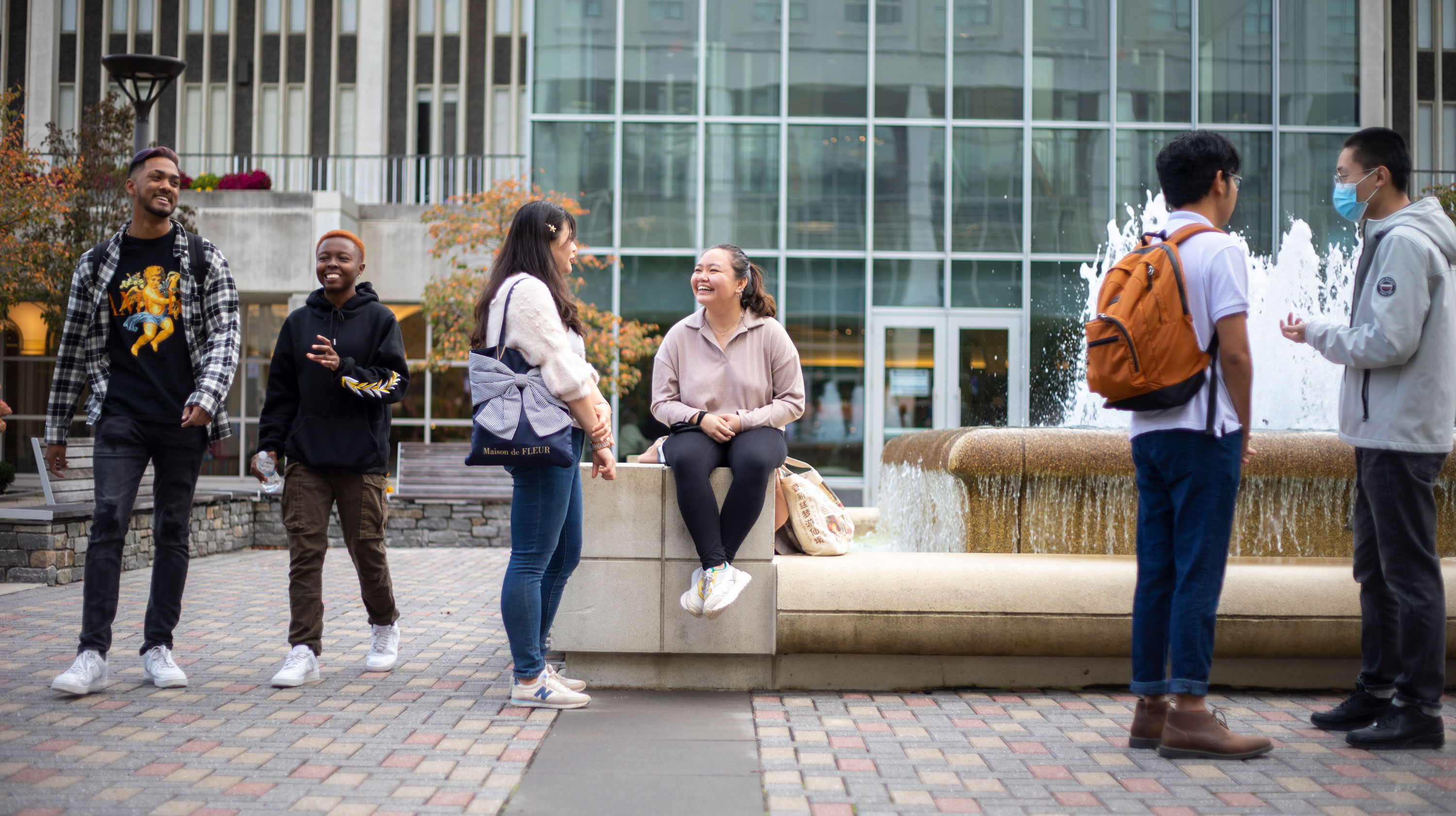 Six students gather outside on campus, speaking and smiling in pairs. Two are walking, three are standing and one is sitting.