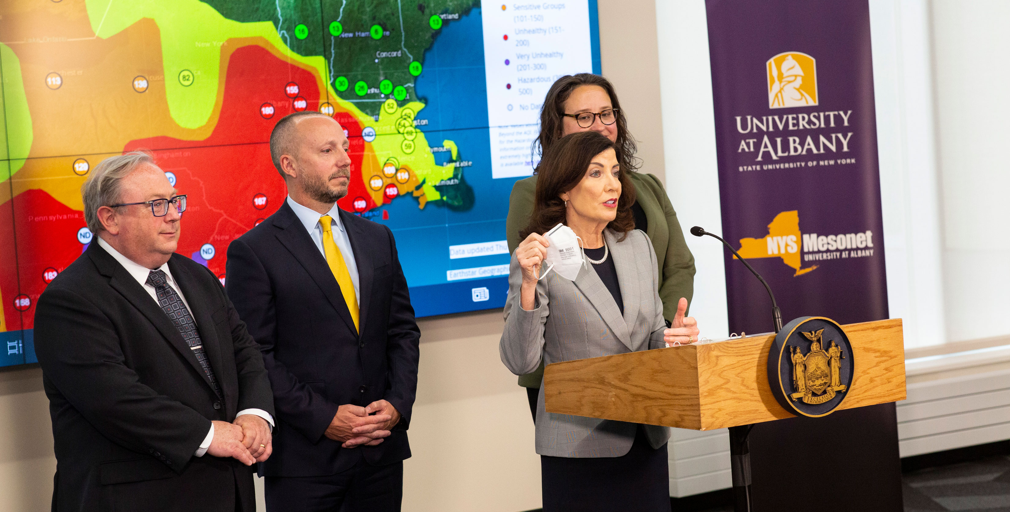 New York State Governor Kathy Hochul holds up a face mask and speaks at a press conference while standing in front of a NYS Mesonet map and UAlbany logo.