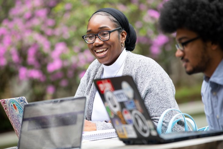 Two smiling students sit at a picnic table with open laptops in the azalea garden.