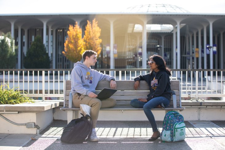 Two students smile as they talk and sit on a bench outside on the Academic Podium at UAlbany.