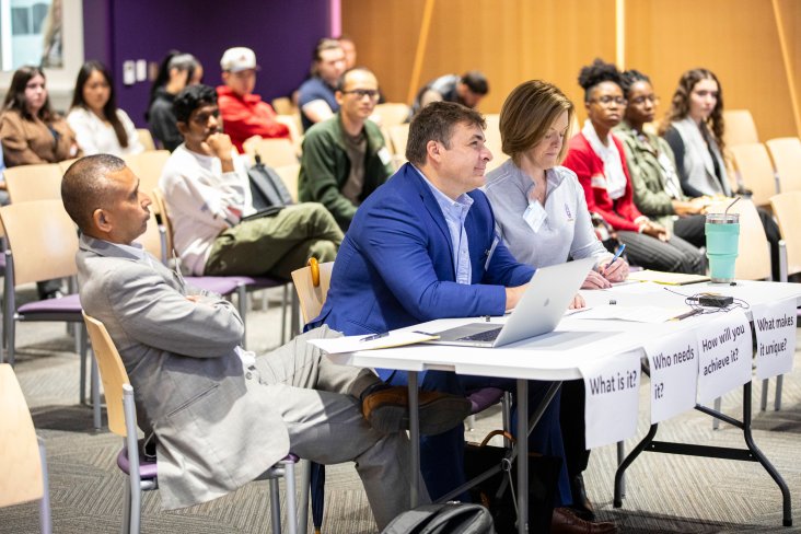A panel of three judges sits at a table in front of an audience at a campus event.