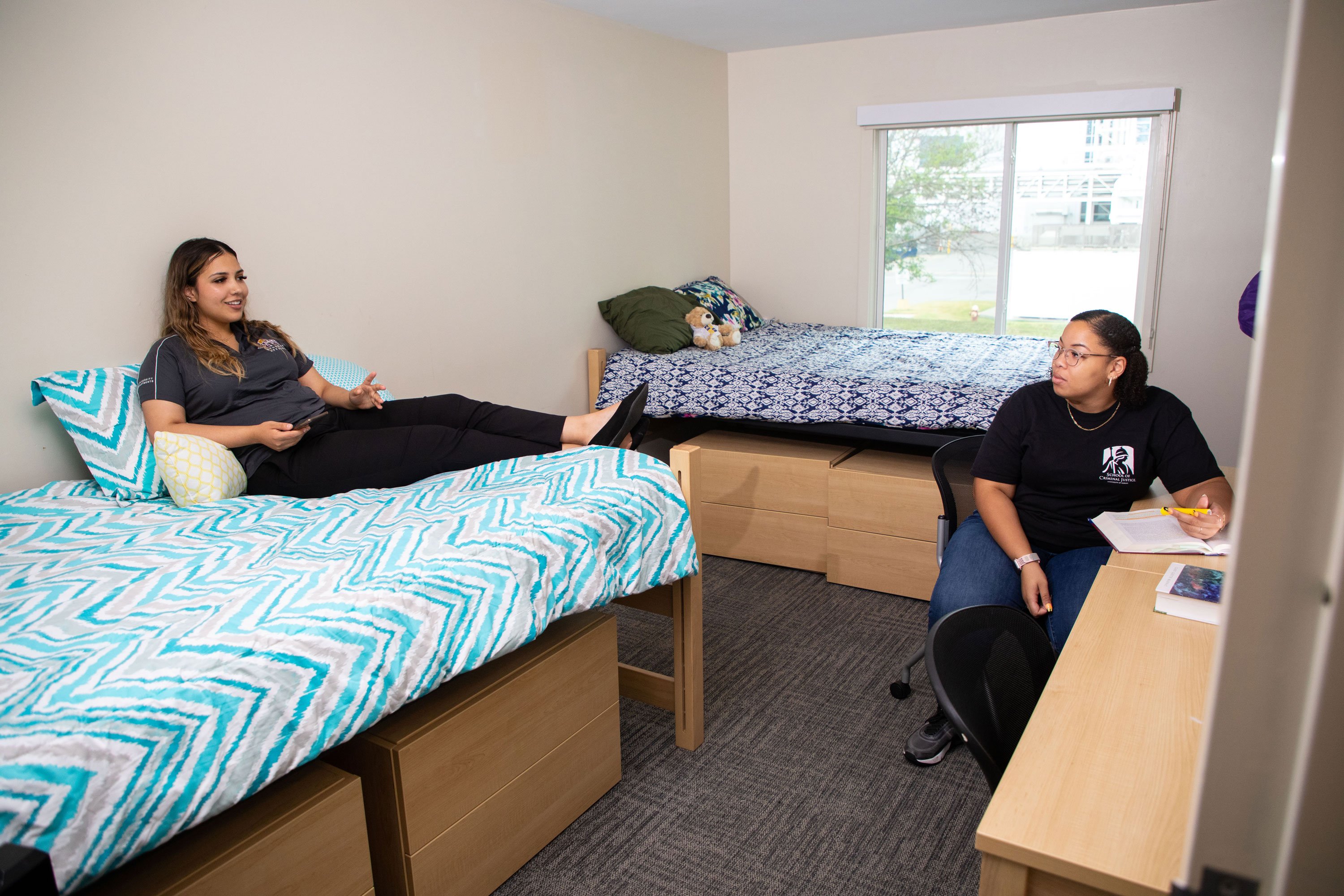 Two students talk inside a double room with two full-sized beds. One student sits on a bed with a phone and another with a text book and highlighter at her desk.