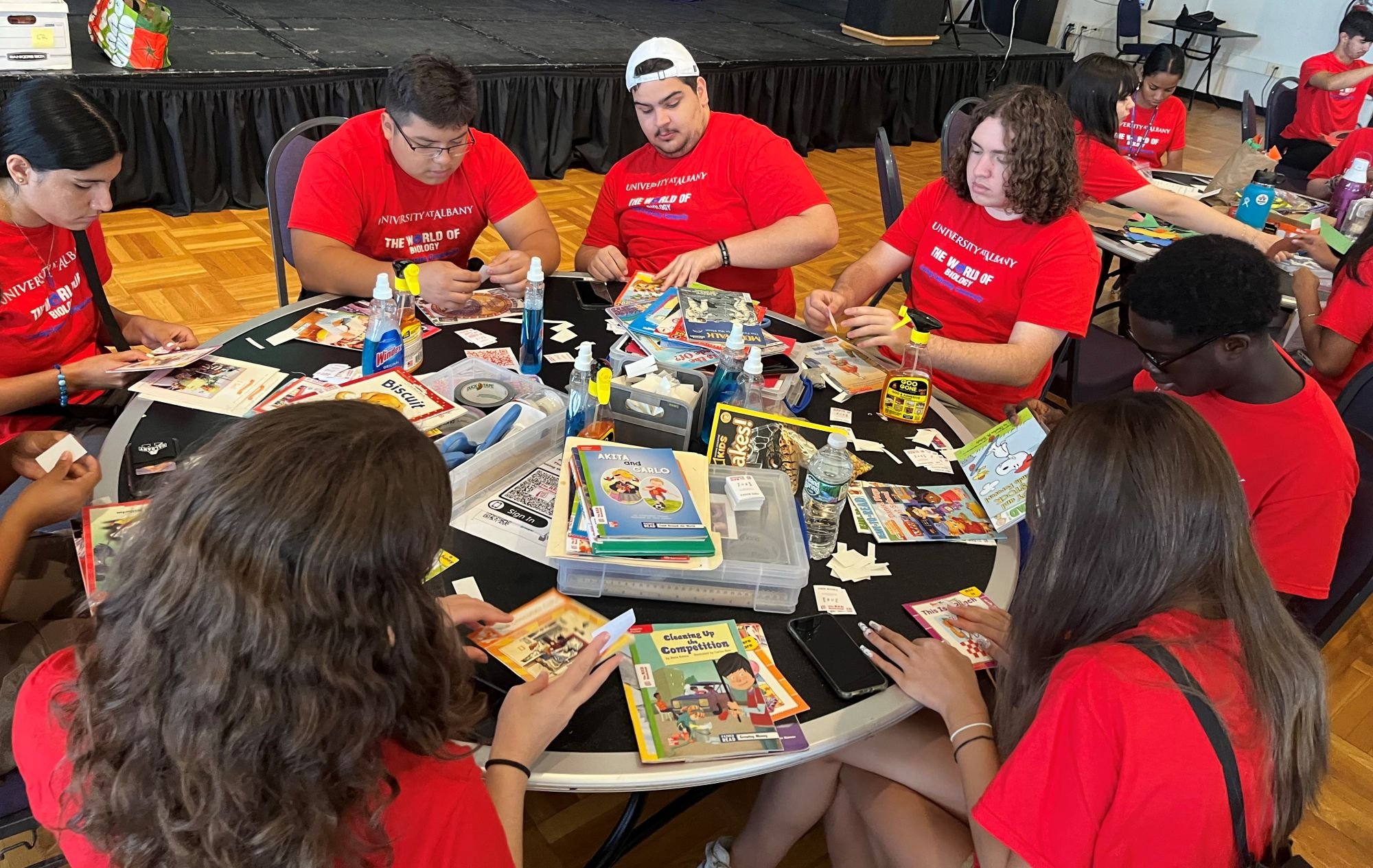 Eight students in matching red T-shirts that say "The World of Biology" are seating around a table and mending used children's books.
