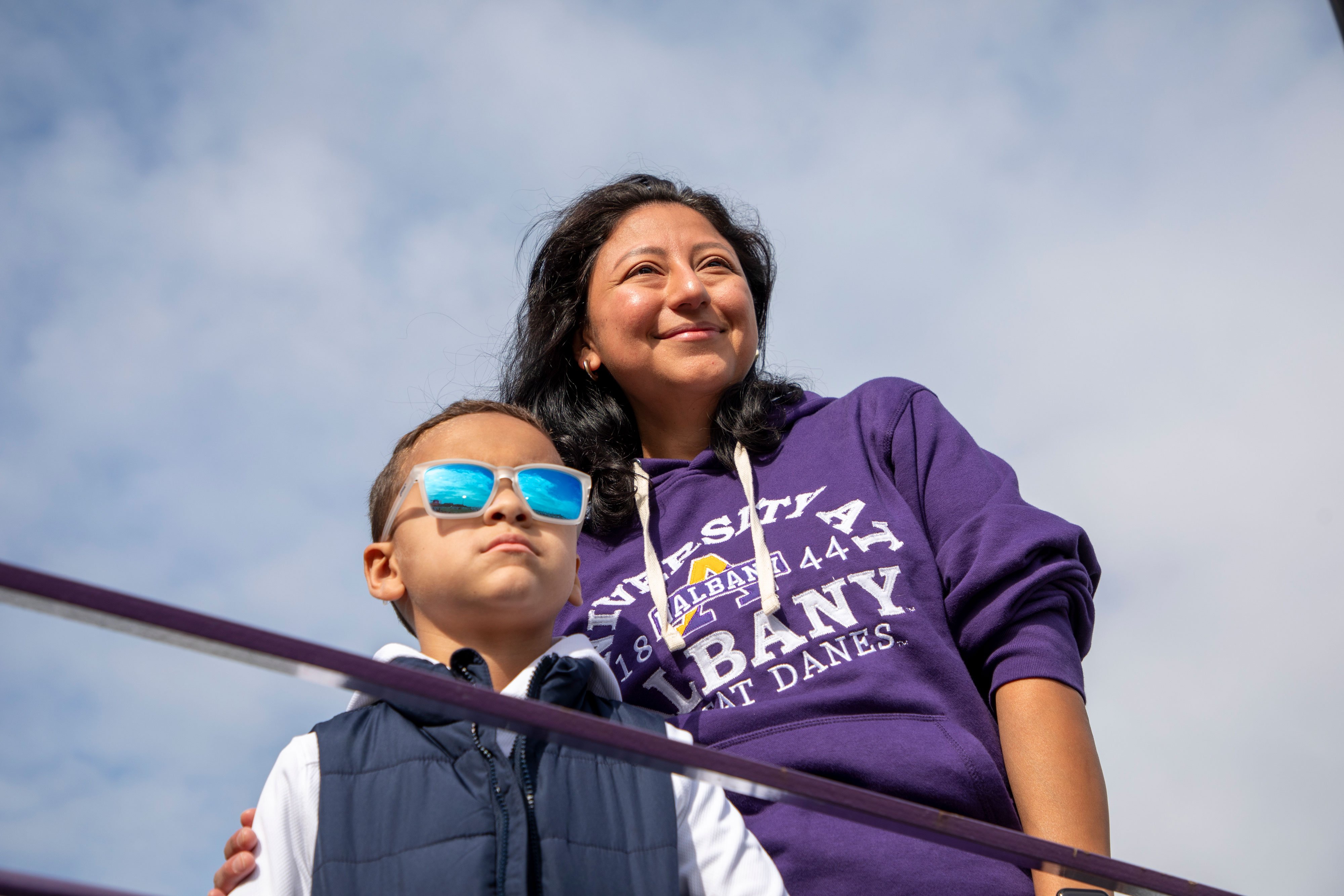 An adult in a UAlbany sweatshirt and a child wearing sunglasses smile as they pose for a photo.