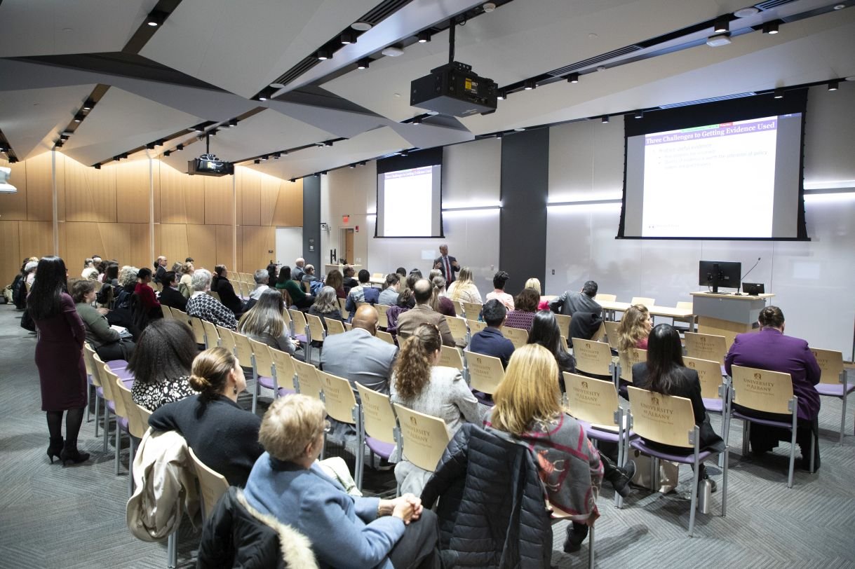 An audience of over 100 people gathered in UAlbany's ETEC building for the ISHE launch event. In this image, all are seated and two projector screens are lit in the background. The visible text reads: "Three Challenges to Getting Evidence Used".