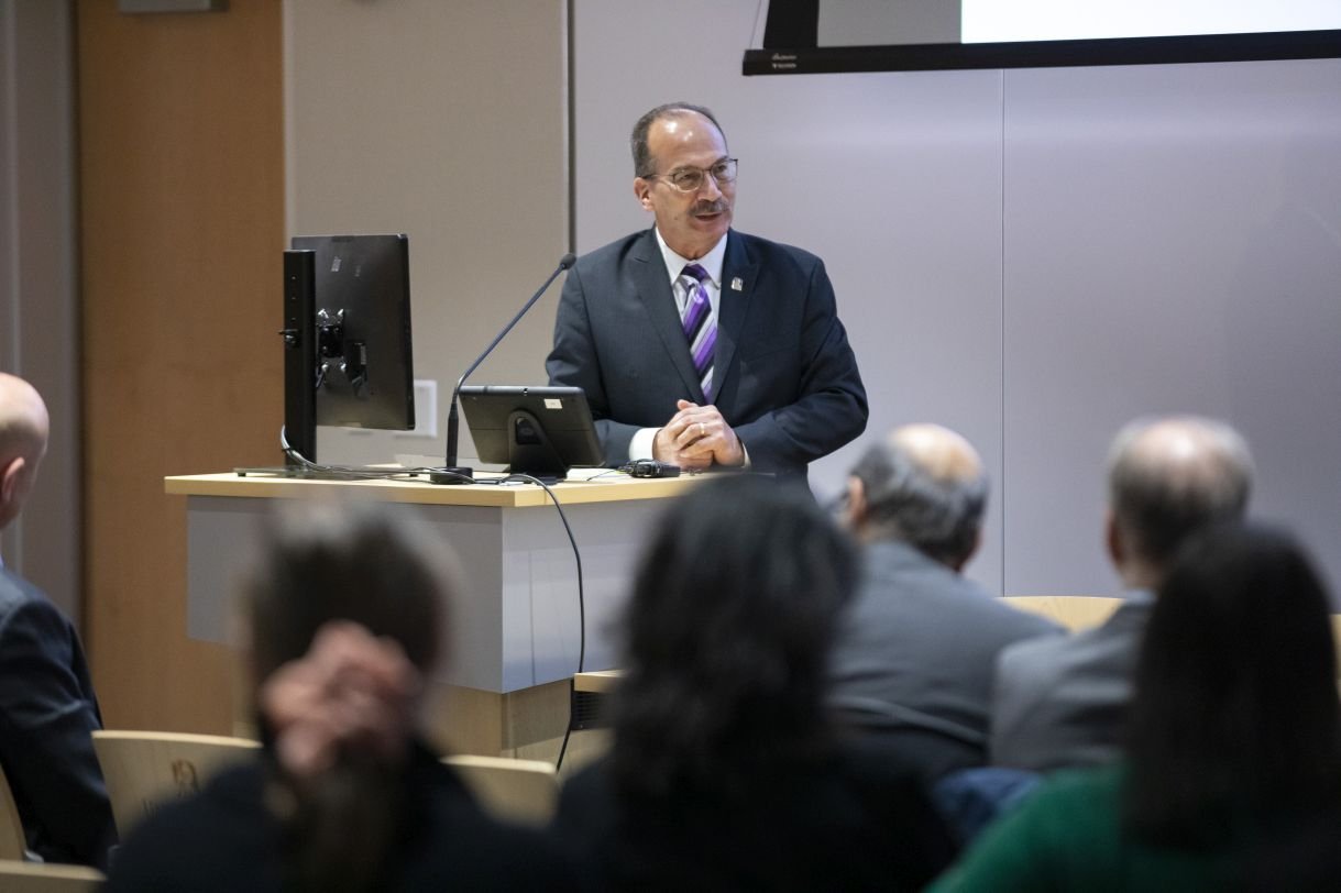 A man wearing glasses, a gray suit, UAlbany lapel pin and purple striped tie stands at a podium in UAlbany's ETEC building to address an audience. 