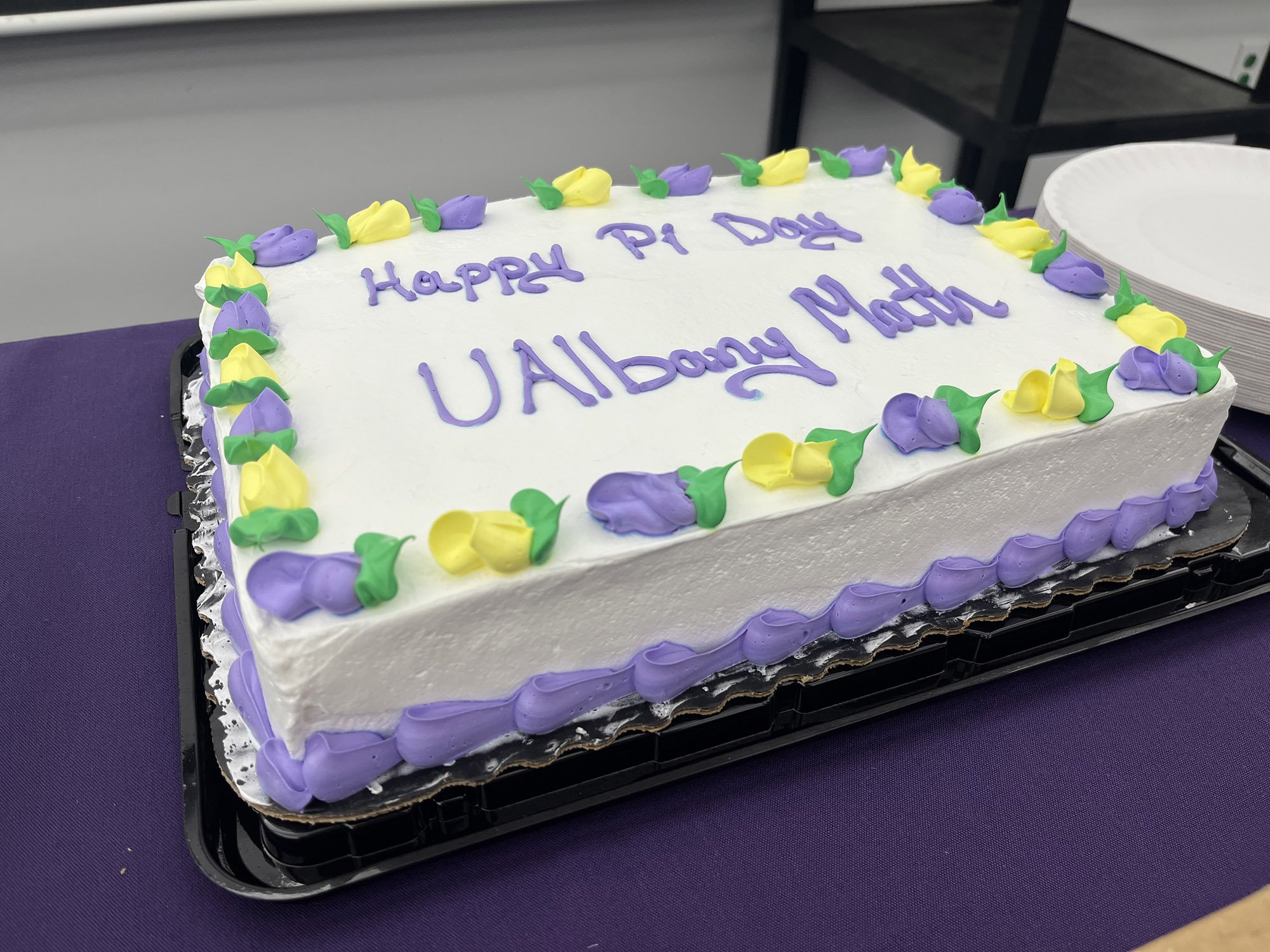 A cake with purple and yellow flowers and purple lettering reads "Happy Pi Day UAlbany Math". 