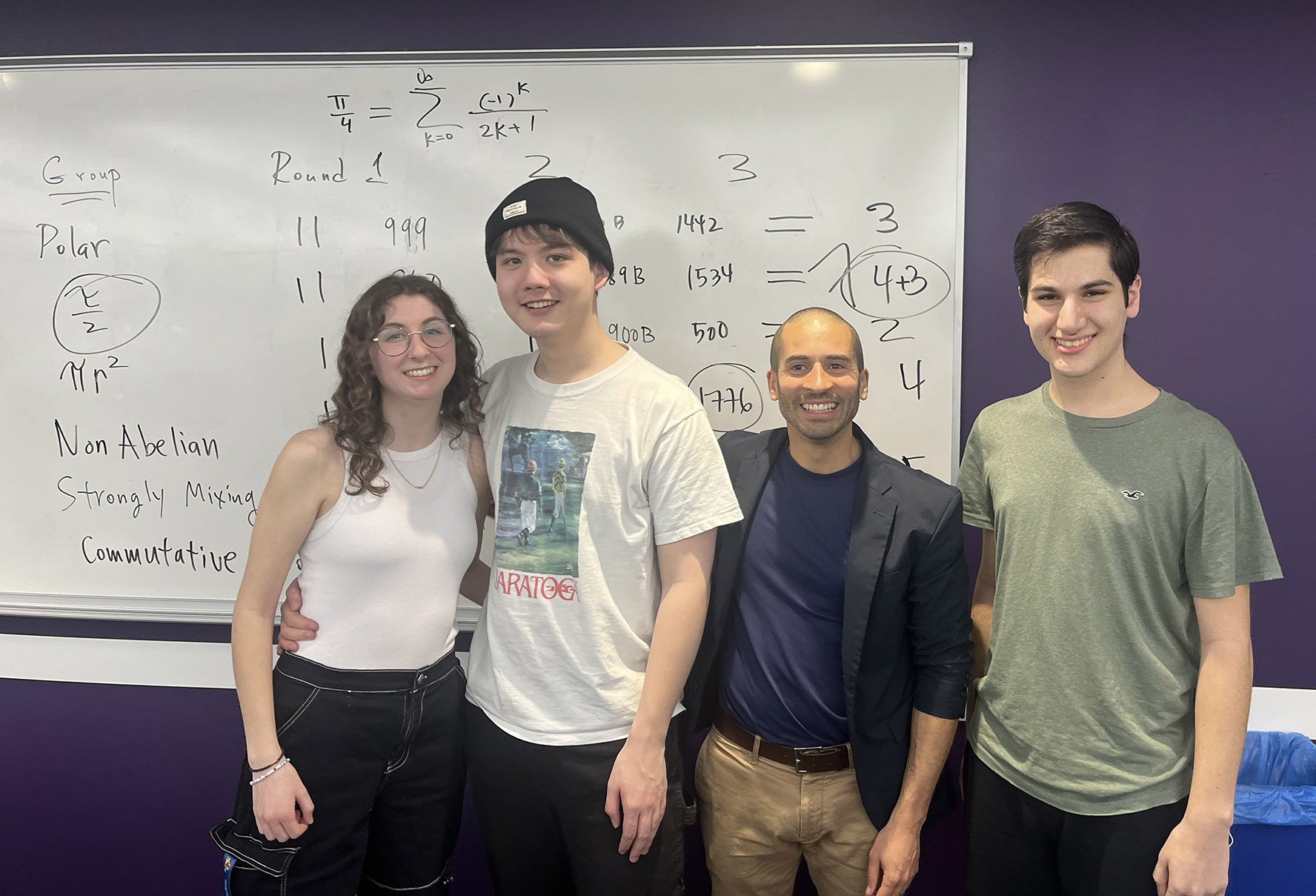 Students and a professor stand in front of a whiteboard with mathematical equations on it.