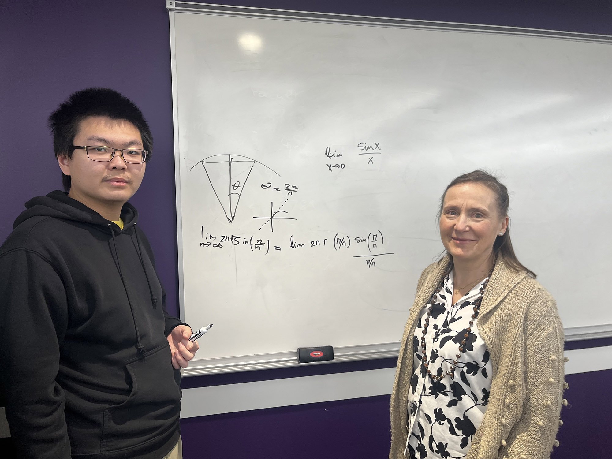 A student and a professor stand in front of a whiteboard with an equation on it.