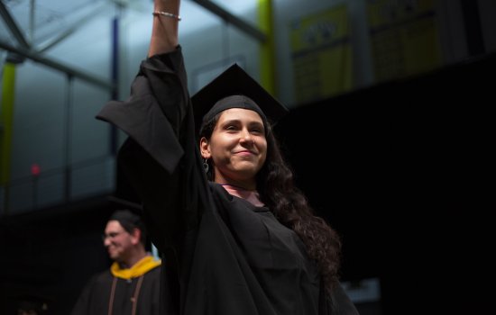 A student smiles and holds up her hand to wave as she crosses the graduation stage.