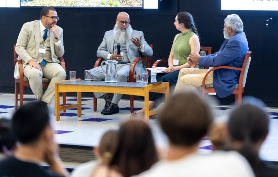 Four people sit around a coffee table set up on a stage. A man with glass and a long beard, wearing a gray suit holds a microphone as he addresses the group.