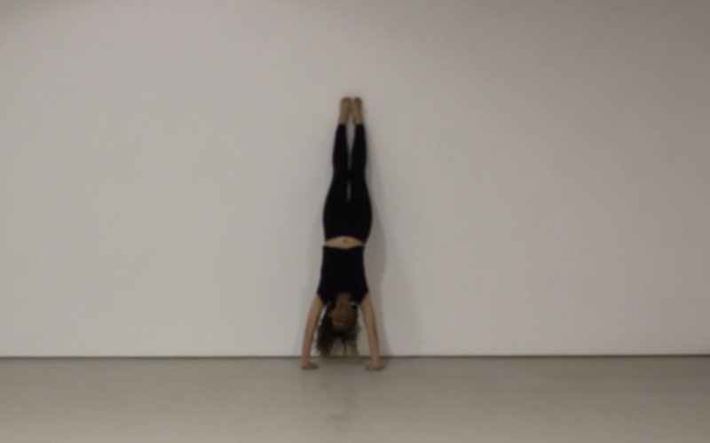 A woman wears all black and does a handstand against a white wall.