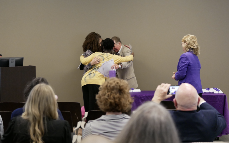 Students huddle around a teacher for a group hug near a purple table as an audience looks on.