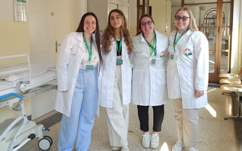 Four female students wearing white medical jackets and green lanyards with nametags stand in a sunlit hospital hallway. All are smiling. Part of a stretcher is visible at left. 