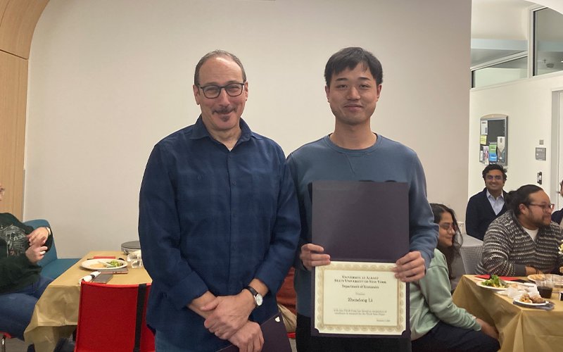 A man wearing a plain slate gray shirt holds a certificate and smiles as he stands on the right side of the image, next to man in a blue plaid button up and glasses.