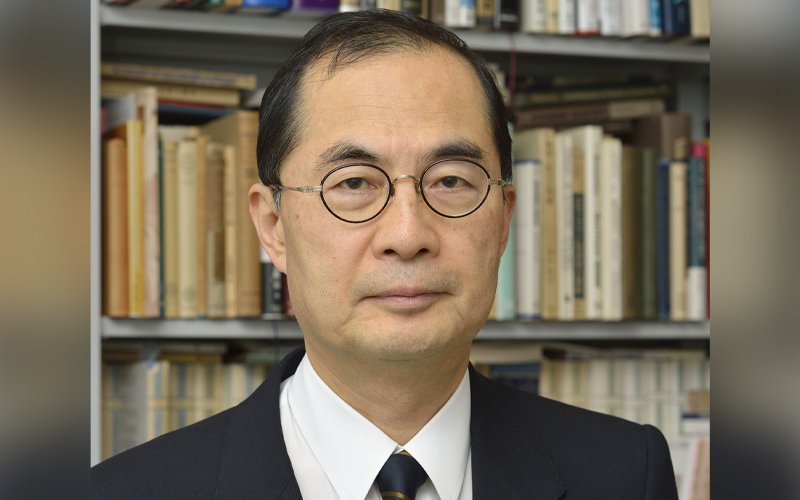 Hiroshi Yoshikawa wears glasses and a suit while standing in front of a bookshelf full of books.