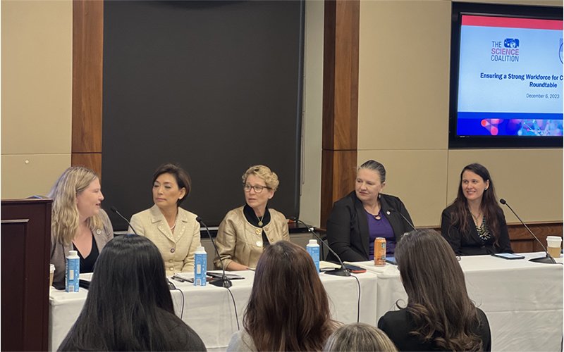 Michele Grimm sits second from left at a table of women dressed in business attire. They are seated at the front of an audience and speaking into table microphones. On the wall behind them, a monitor reads, "The Science Coalition/Ensuring a Strong Workforce for... Roundtable" with some words cut out of the frame.