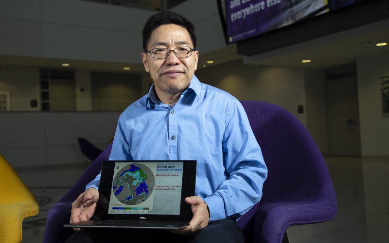 Aiguo Dai holds a laptop with a globe graphic displayed inside University Hall.