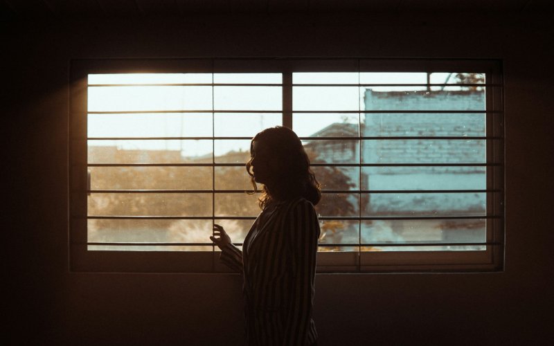 The silhouette of a woman is seen standing in front of a window with horizontal metal bars on it.