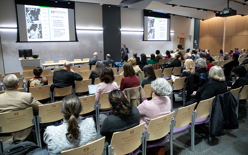 Adam Gamoran wears a dark suit and red tie as he stands in front of a crowded room in UAlbany's ETEC building during the Inaugural Speaker Series and Institute for Social and Health Equity Launch. His lecture slides, too small to be readable in the photo, are projected on screens behind him.