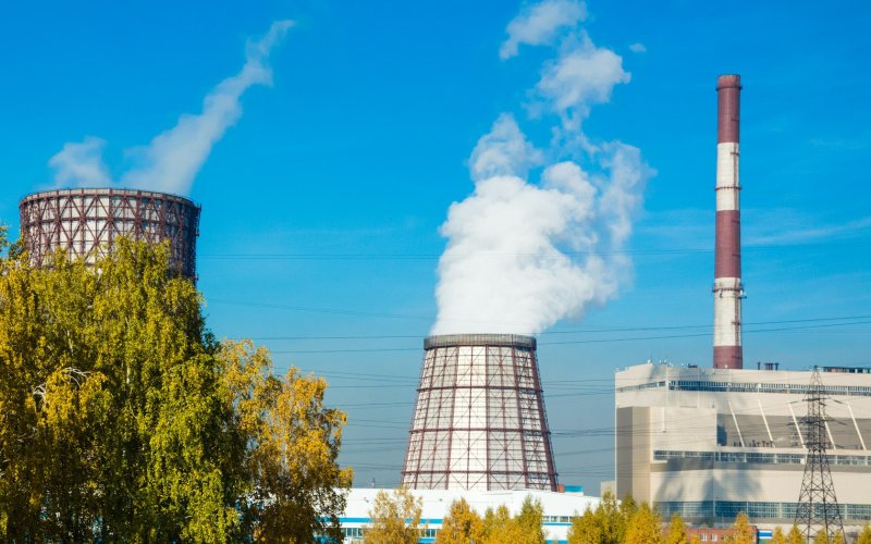 Landscape image featuring two large industrial cooling towers near large nondescript buildings with powerlines and a tall smokestack. Trees are in the foreground and plumes of mist from the towers drift skyward.