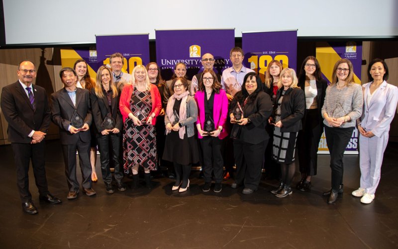 The president and provost flank a group of 16 people standing in two rows, smiling and holding tear-shaped glass awards.