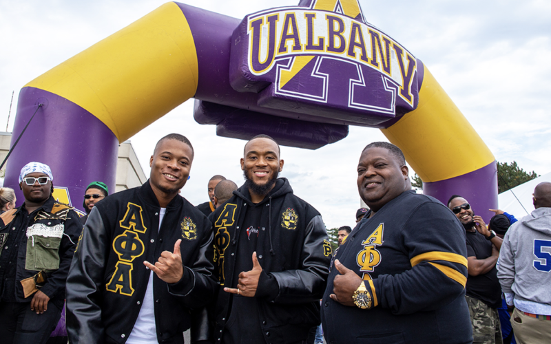 Three men in black and gold Alpha Phi Alpha fraternity jackets smile and flash shaka signs in front of a large inflated purple and gold UAlbany arch. 