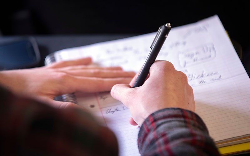 Viewed from over a student's shoulder, two hands write in a notebook with a pen. The student is wearing a long-sleeved flannel shirt.