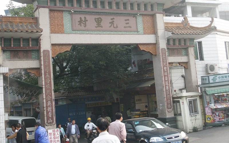 A clay and green-brick archway hangs over a street in China, with deep brown Chinese lettering. Below, people and cars travel through the archway.