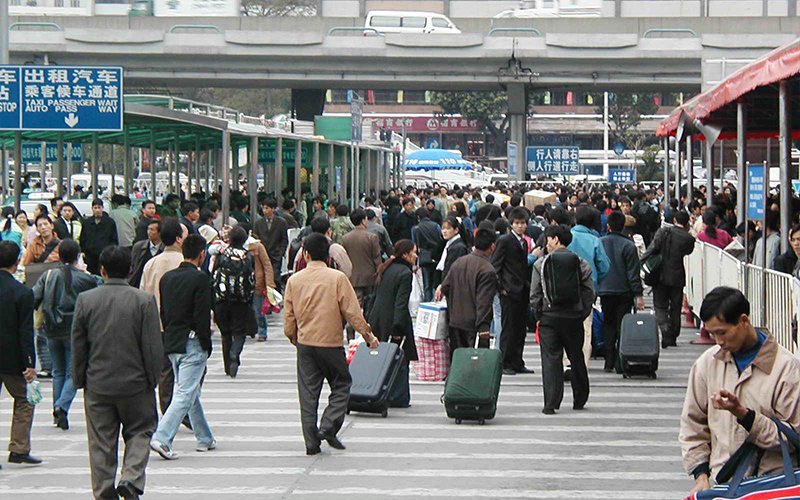 People walk in a crowded crosswalk in China, many rolling suitcases behind them. Traffic signs and metal partitions surround the crosswalk.