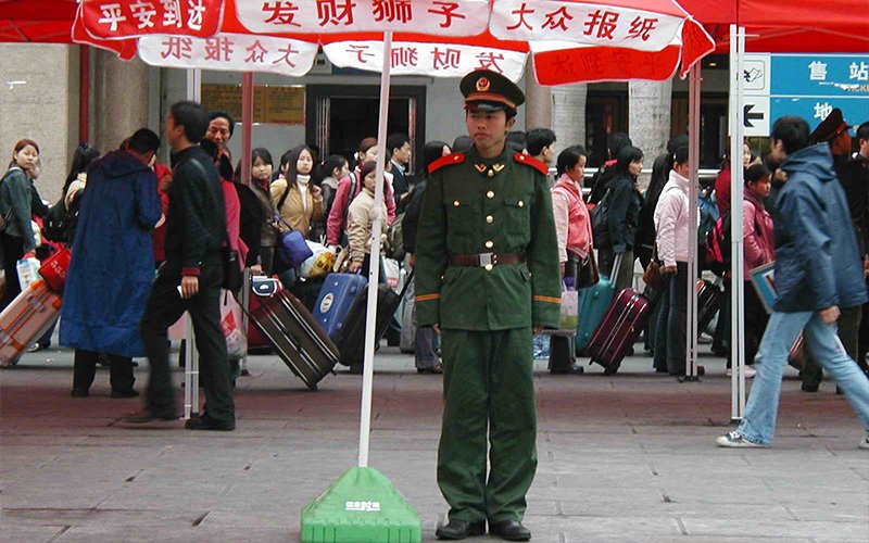 A man in a green and red military uniform stands below a red and white umbrella. Behind him, a crowded line of people wait with suitcases in hand.