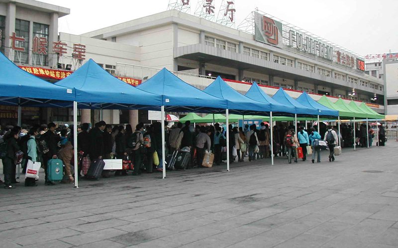 People stand beneath a row of teal pop-up tents in front of Guangzhou Station in China. Behind them, the building displays signs written in Chinese, and the logo for Fujifilm, and a digital clock face lit up in red.