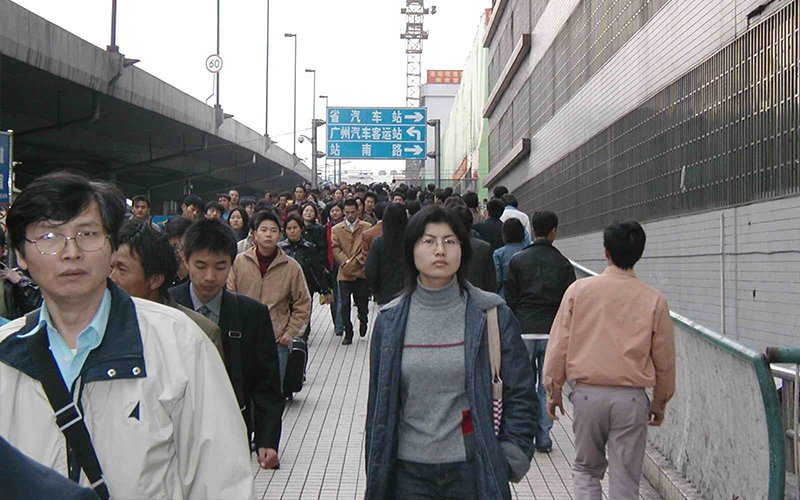 A woman stands on a walkway in china, surrounded by a sea of people walking by. Behind her, a traffic sign displays Chinese text that points to three locations. The text translates to "Provincial bus station", "Guangzhou Bus Terminal," and "Zhannan Road".