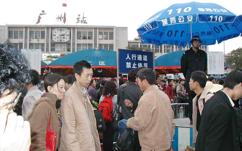 A crowd of people walk in front of Guangzhou Station in China. The station has red and teal awnings out front, a clock on its facade and a sign in Chinese that translate to, "Guangzhou Station."