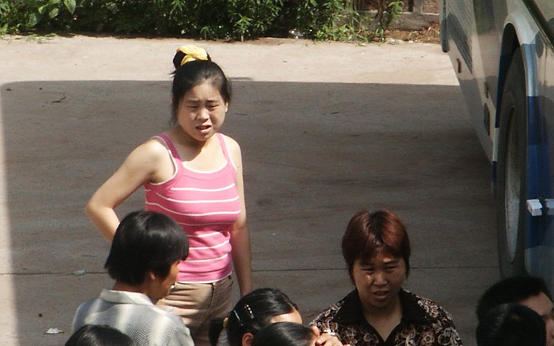 A Chinese woman wearing a pink and white striped tank top stands on the pavement, with a crowd of people, next to a shuttlebus.