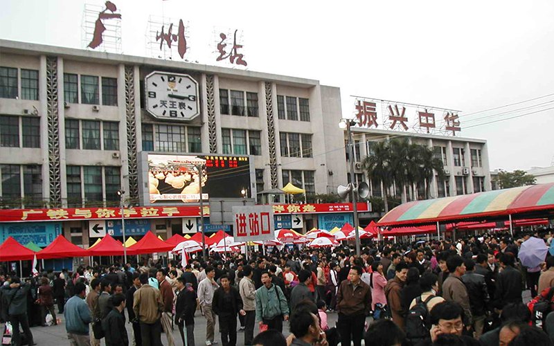 A crowd of people walk in front of Guangzhou Station in China. The station has red tents and red and teal awnings out front, a clock on its facade and signs in Chinese that translate to, "Guangzhou Station" and "Revitalize China."