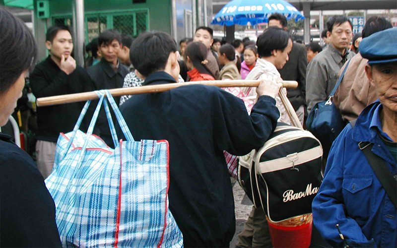 A man walks through a crowd in China, using a wooden pole to carry two large bags slung over his shoulder. One is a light-blue plaid pattern and the other is black with white trim and white text that reads "BaoMa."